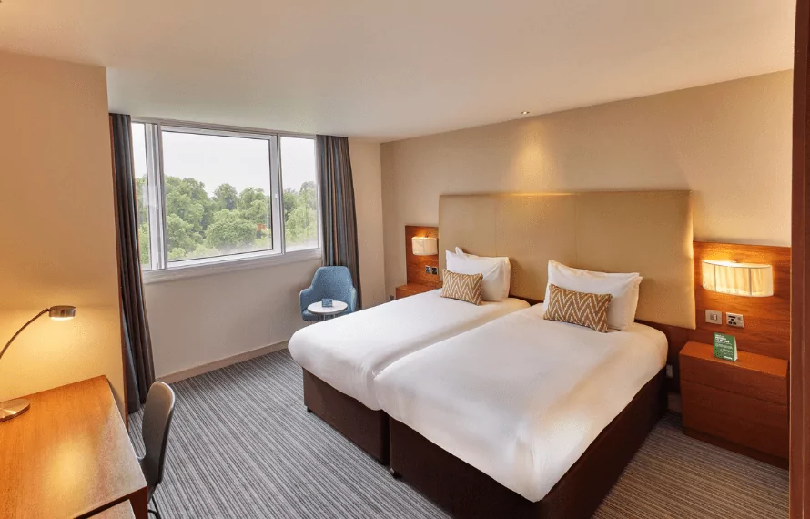 Hotel room with two single beds, zigzag cushions, nightstands, and a blue chair by the window