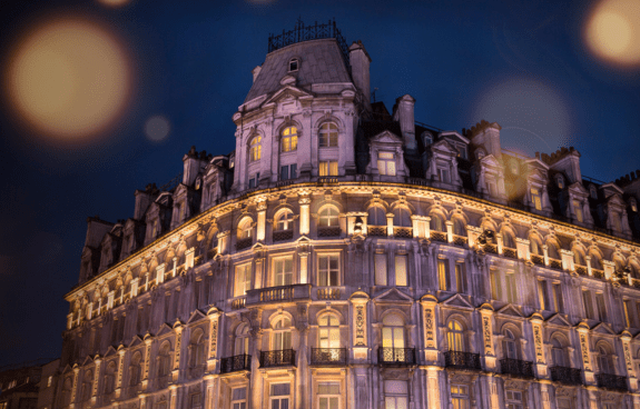 Ornate multi-story building illuminated at night with decorative architecture and light flares in the dark blue sky
