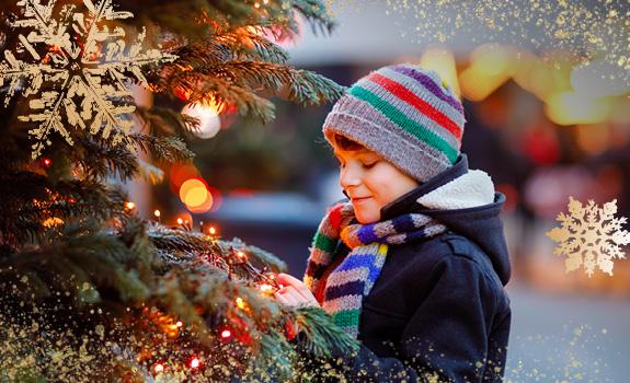 Child in festive winter wear beside a decorated Christmas tree with glowing lights