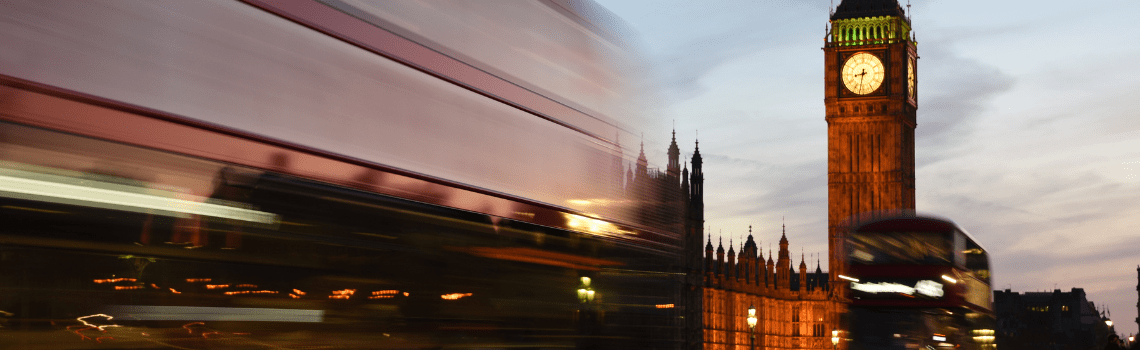 Motion‑blurred red double‑decker buses pass Big Ben and the Houses of Parliament at dusk, with the illuminated clock tower standing against a blue evening sky.