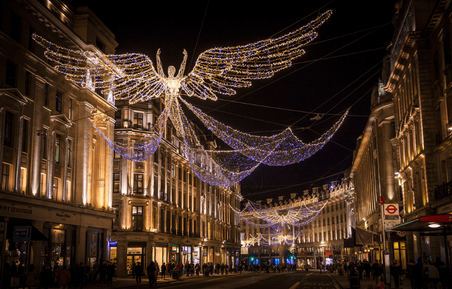 Nighttime street scene with a large angel-shaped light installation above, surrounded by festive string lights and warmly lit buildings