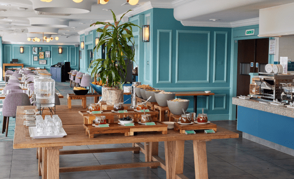 Buffet setup in a bright dining area with wooden tables displaying glass pitchers, bowls of snacks, and tea jars, with seating in the background