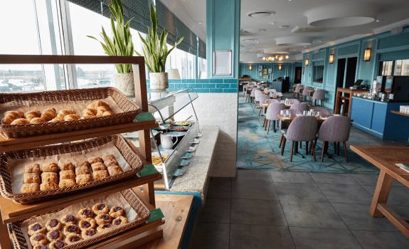 Breakfast buffet in a bright dining area with baskets of pastries, a counter with condiments, and tables with chairs arranged in the background.