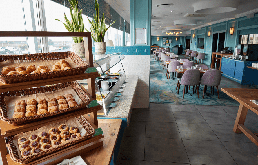 Breakfast buffet in a bright dining area with baskets of pastries, a counter with condiments, and tables with chairs arranged in the background.