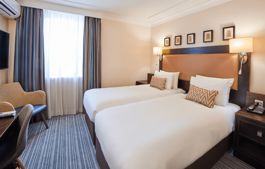 Hotel room with two single beds, white bedding, patterned accent pillows, a large padded headboard, bedside lamps, a desk with a chair, and a window with curtains