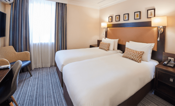 Hotel room with two single beds, white bedding, patterned accent pillows, a large padded headboard, bedside lamps, a desk with a chair, and a window with curtains