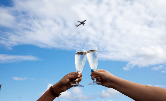 Two hands holding champagne glasses in a toast against a blue sky with clouds and an airplane flying overhead.