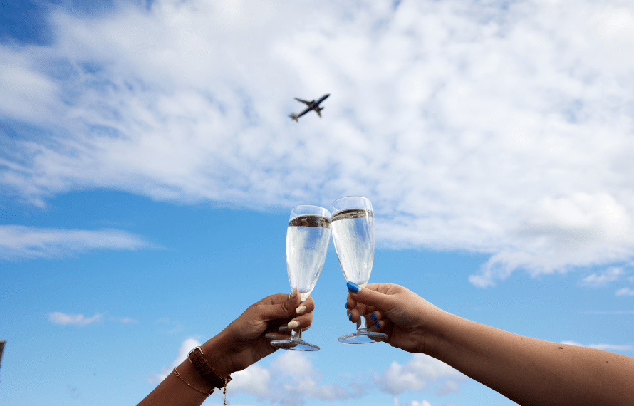 Two hands holding champagne glasses in a toast against a blue sky with clouds and an airplane flying overhead.