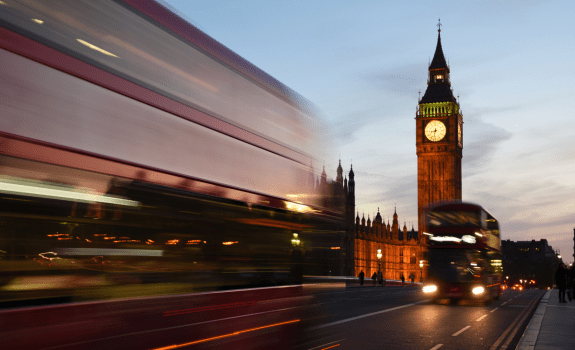 Motion‑blurred red double‑decker buses pass Big Ben and the Houses of Parliament at dusk, with the illuminated clock tower standing against a blue evening sky.