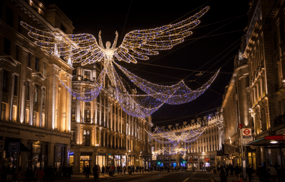 Nighttime street scene with a large angel-shaped light installation above, surrounded by festive string lights and warmly lit buildings