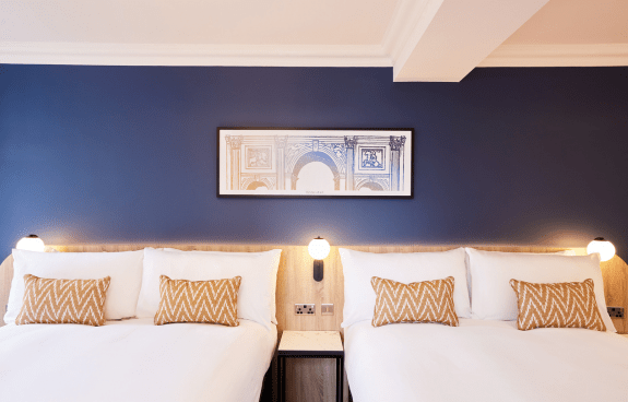 Close‑up of king beds with white linens, patterned cushions, and bedside lighting against a blue wall.