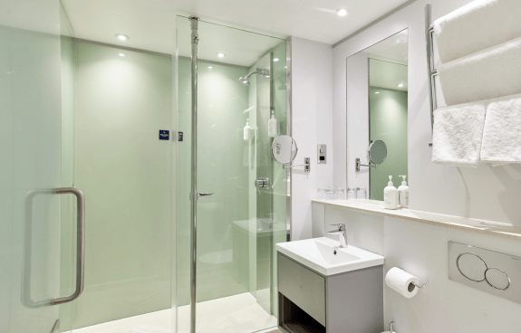 Modern hotel bathroom featuring a glass walk‑in shower, compact sink, wall mirror, and white towels.