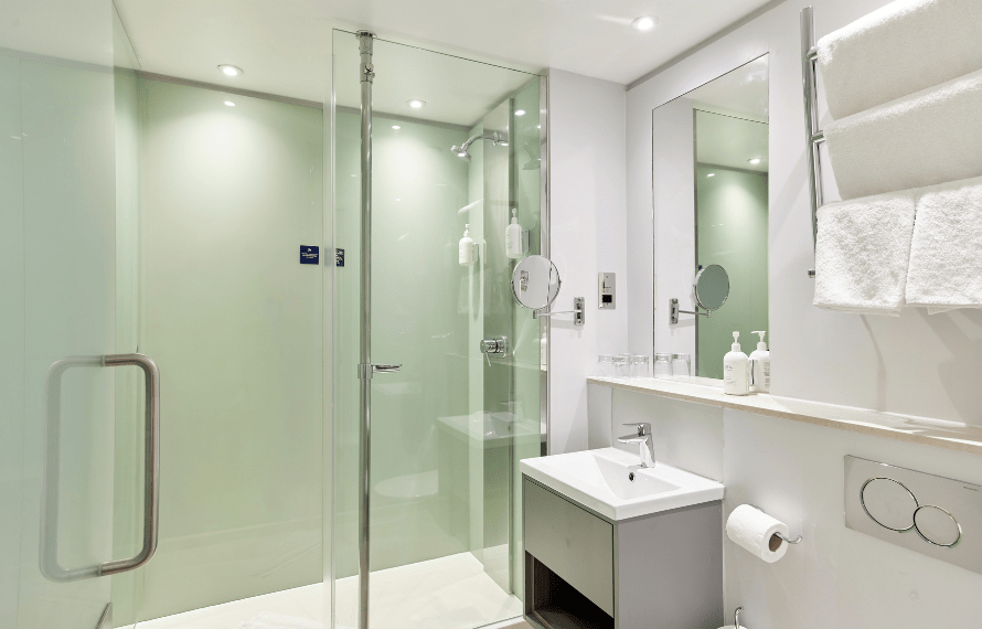 Modern hotel bathroom featuring a glass walk‑in shower, compact sink, wall mirror, and white towels.