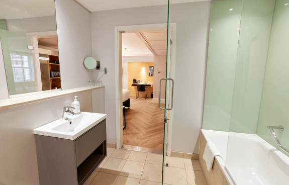 Spacious hotel bathroom with glass shower, bathtub, vanity sink, and tiled flooring leading into the bedroom.