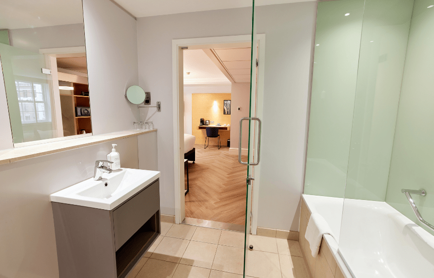 Spacious hotel bathroom with glass shower, bathtub, vanity sink, and tiled flooring leading into the bedroom.