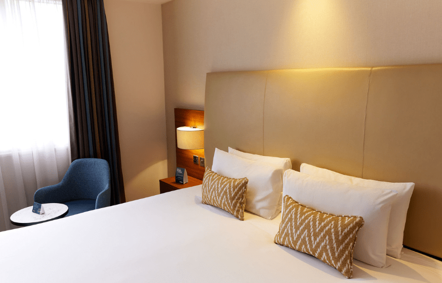 A neatly made bed, zigzag-patterned pillows, nightstand, and a blue armchair near the window