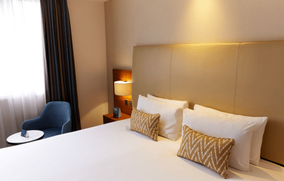 A neatly made bed, zigzag-patterned pillows, nightstand, and a blue armchair near the window