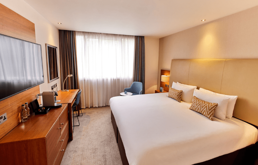 Hotel room with a large bed, desk with coffee setup, armchair, and wall-mounted TV