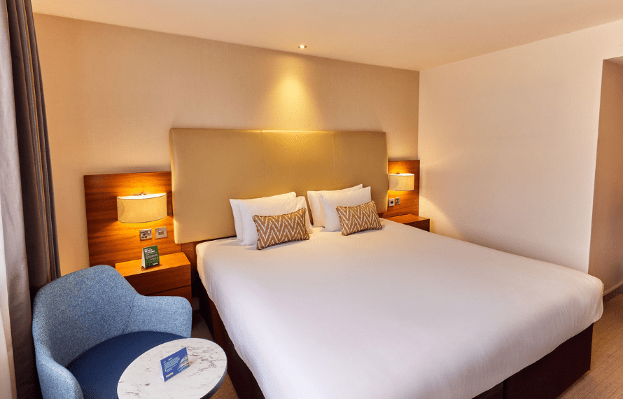 Deluxe king room with large bed, patterned pillows, nightstands, and a blue chair beside a marble-topped table