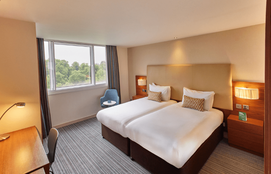 Hotel room with two single beds, zigzag cushions, nightstands, and a blue chair by the window