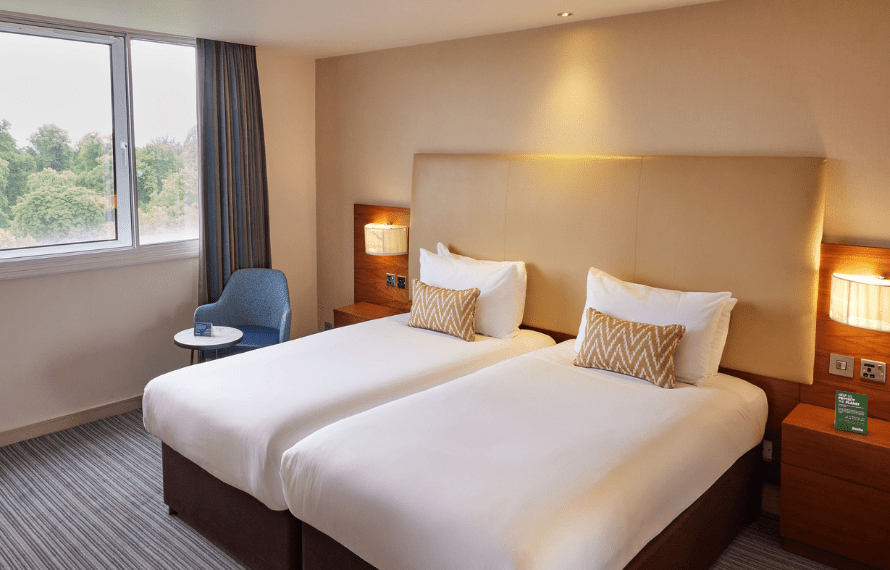 Hotel room with two single beds, zigzag cushions, nightstands, and a blue chair by the window