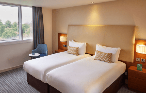 Hotel room with two single beds, zigzag cushions, nightstands, and a blue chair by the window