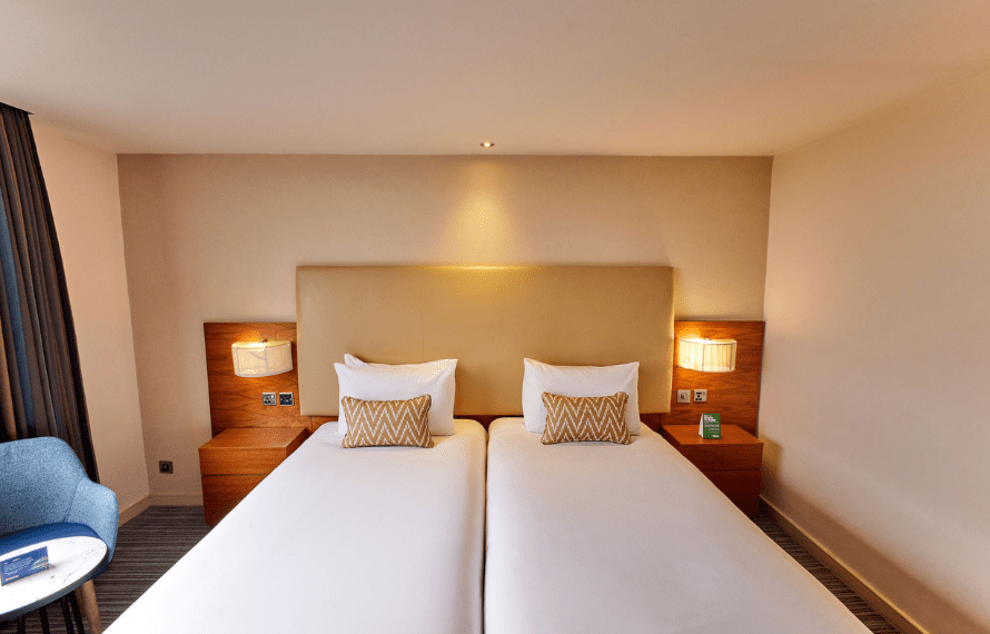 Hotel room with two single beds, zigzag-patterned pillows, nightstands, and a blue chair beside a small table