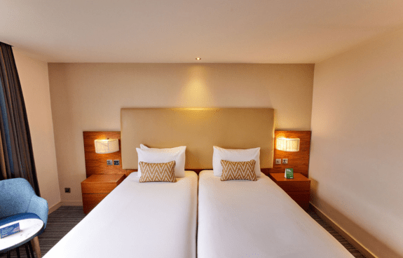 Hotel room with two single beds, zigzag-patterned pillows, nightstands, and a blue chair beside a small table