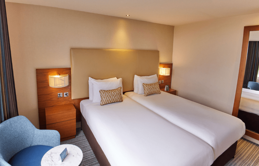 Hotel room with two single beds, nightstands, a blue chair, and a wall-mounted mirror