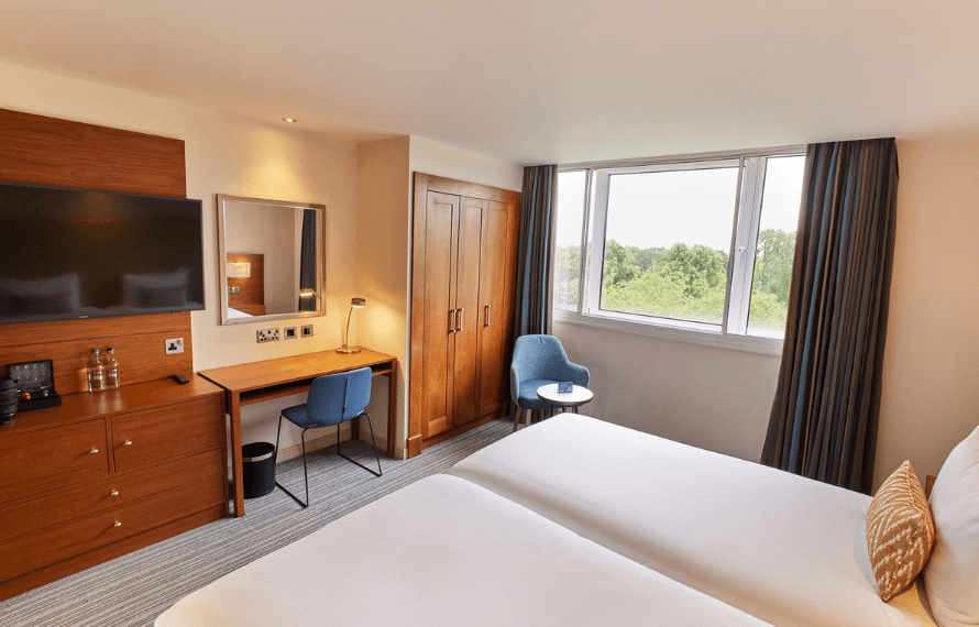 Hotel room with two beds, desk and TV, armchair by the window, and a view of greenery outside