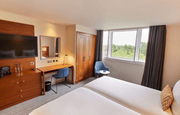 Hotel room with two beds, desk and TV, armchair by the window, and a view of greenery outside