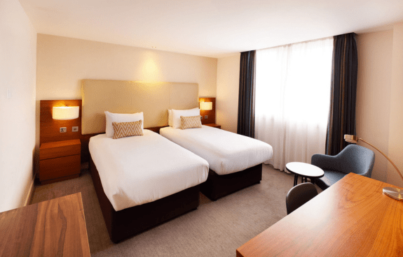 Hotel room with two single beds, bedside tables, armchair, desk, and a large window with curtains