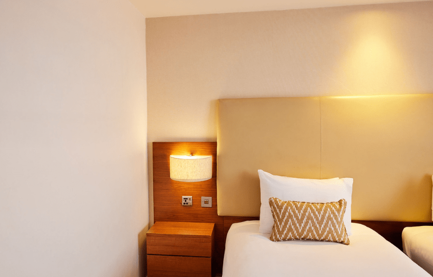 Bed with white sheets, zigzag pillow, beige headboard, and a lit lamp on a wooden nightstand