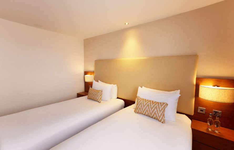 Hotel room with two single beds, zigzag-patterned pillows, nightstands with lamps, and bottled water