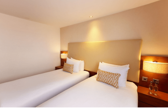 Hotel room with two single beds, zigzag-patterned pillows, nightstands with lamps, and bottled water